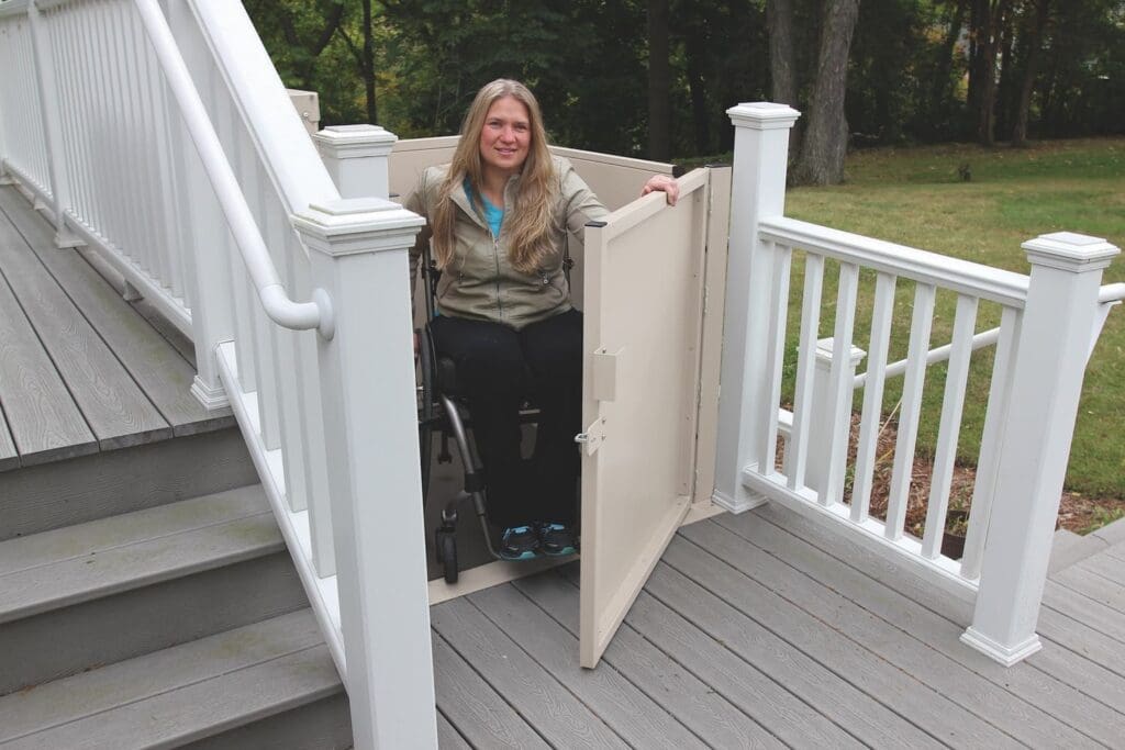 A woman in a wheelchair uses a Bruno VPL-3100/3200 Series Vertical Platform Lift beside an outdoor staircase on a wooden deck with white railings, surrounded by trees.
