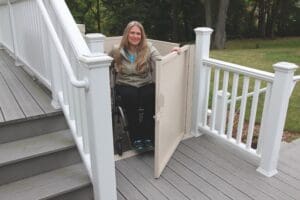 A woman in a wheelchair uses a Bruno VPL-3100/3200 Series Vertical Platform Lift beside an outdoor staircase on a wooden deck with white railings, surrounded by trees.