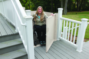 A woman in a wheelchair uses a Bruno VPL - 3100/3200 Series Vertical Platform Lift to reach a raised deck next to stairs, with white railings and greenery in the background.