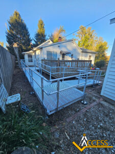 A modular metal wheelchair ramp with railings leads up to the porch of a white house. Trees surround the area and the Access company logo is in the bottom right corner. It is a sunny day.