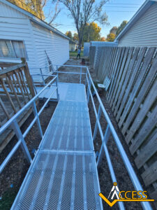 A metal wheelchair ramp leads from a wooden porch to a gravel path beside a house and a wooden fence. The ACCESS logo appears in the lower right corner. Trees and a shed are visible in the background.