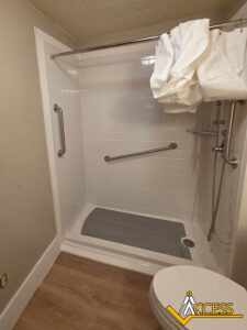 Accessible walk-in shower with white tile, grab bars on two walls, a handheld showerhead, a gray non-slip mat, and a white shower curtain pulled to one side. Wooden floor outside and ACCESS logo in the corner.