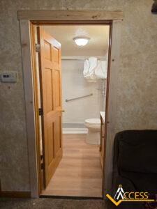Open wooden door revealing a small, accessible bathroom with a toilet, grab bar, shower, and sink; beige walls and wooden floor; Access logo in lower right corner.