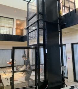 A person stands near a sleek black V-1504 Enclosed Vertical Platform Lift by Savaria with glass doors on the ground floor of a multi-story building with open interiors, visible upper-level railings, and construction materials around.