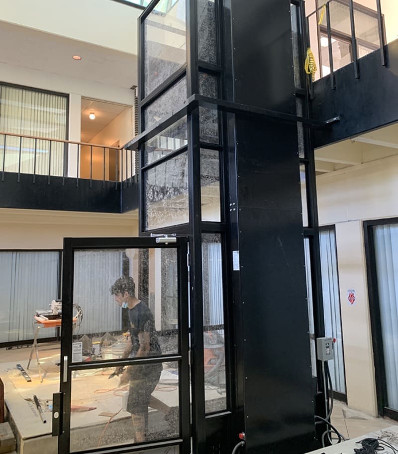 A person stands near a sleek black V-1504 Enclosed Vertical Platform Lift by Savaria with glass doors on the ground floor of a multi-story building with open interiors, visible upper-level railings, and construction materials around. A person stands near a sleek black V-1504 Enclosed Vertical Platform Lift by Savaria with glass doors on the ground floor of a multi-story building with open interiors, visible upper-level railings, and construction materials around.