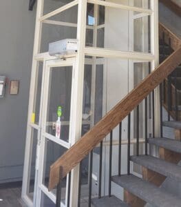 A V-1504 Enclosed Vertical Platform Lift by Savaria is installed beside a wooden staircase with black railings in a modern building. The compact glass-walled lift includes a control panel and safety instructions on its door.
