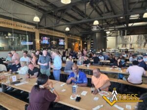 A large group of people sit and stand around long wooden tables in a busy, industrial-style brewery or restaurant, embodying innovation and community. Many are eating, drinking, and socializing. The Access logo is in the bottom-right corner.