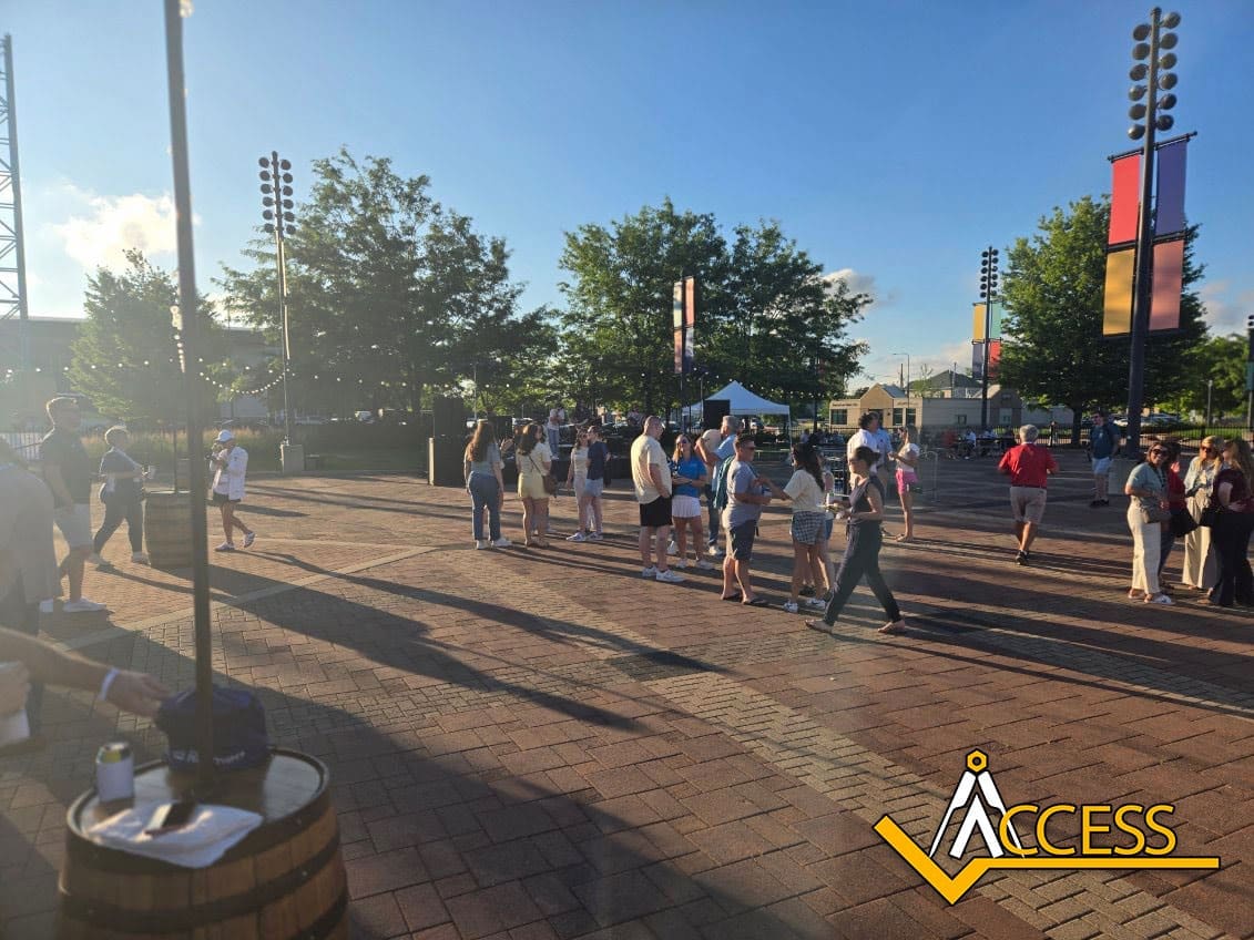 People gather outdoors on a sunny day in a plaza with trees and banners, some standing in groups while others walk. The event celebrates industry leadership at the VGM Heartland Conference, with a yellow “ACCESS” logo visible on the brick ground.