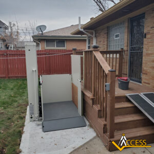 A vertical platform lift is installed beside outdoor wooden stairs, providing wheelchair access to the back door of a brick house with a red fenced yard. The Access company logo is visible in the lower right corner.