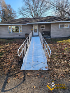 A metal wheelchair ramp with handrails leads to the front door of a single-story house. Fallen autumn leaves cover the ground, and the ACCESS logo appears in the lower right corner.