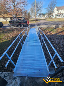 A metal wheelchair ramp with railings leads down to a residential street. Fallen leaves are scattered on the ground, and a trailer is parked nearby. The ACESS logo is visible in the lower right corner.