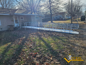 A metal wheelchair ramp with railings leads from the driveway to the front door of a single-story house. Fallen leaves cover the ground and trees without leaves are in the yard. The Access company logo is at the bottom right corner.