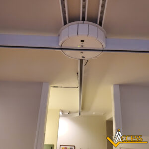 Ceiling-mounted motorized track system with a circular motor unit and curved rail, installed above a doorway in an indoor setting. The Access logo appears in the bottom right corner.
