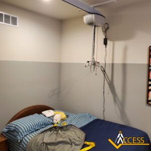 A bedroom with a bed, striped bedding, and stuffed toys, featuring a ceiling-mounted patient lift system above the bed. The Access company logo is in the bottom right corner.