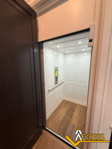 A modern residential elevator with white paneled walls, wood floor, and overhead lighting is open. There is a control panel inside and a dark door frame. The Access logo appears in the bottom right corner.