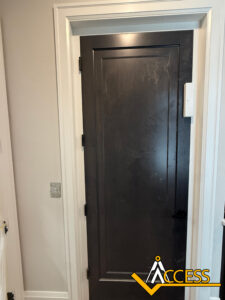A closed dark wooden door with a black handle, set in a white frame. A light switch is on the left wall, and a small rectangular device is mounted on the upper right side of the door frame. ACCESS logo at bottom right.