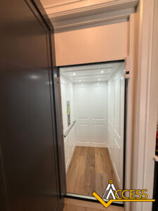 A residential elevator with white paneled walls, wood flooring, built-in lighting, and a control panel inside. The elevator door is open, and the Access logo is visible in the bottom right corner.