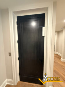 A dark wooden door in a white frame with a rectangular device mounted on the wall next to it. The floor is light wood, and the ACCESS logo is in the bottom right corner.