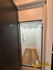 A modern residential elevator with white paneled walls, wooden flooring, and ceiling lights. The elevator door is open, and a control panel is visible on the left wall. The Access company logo is in the bottom right corner.