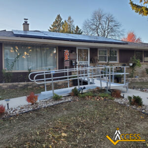 A metal wheelchair ramp leads to the front door of a one-story house with solar panels on the roof. The yard has small shrubs and autumn trees. The Access logo is in the bottom right corner.
