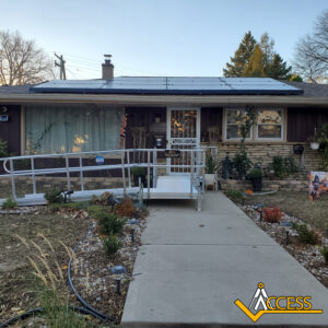 A single-story house with solar panels on the roof, a metal wheelchair ramp leading to the front door, and some plants in the yard. The Access logo is in the bottom right corner.