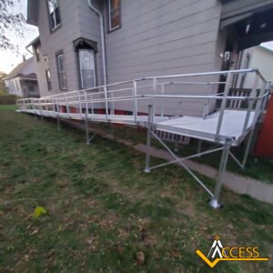 A long, silver metal wheelchair ramp with railings is installed alongside a house, providing accessible entry. The ramp extends over a grassy slope and connects to the porch. The ACCESS logo appears in the lower-right corner.