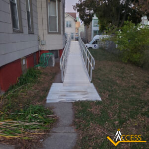 A metal wheelchair ramp with handrails leads from a sidewalk to the side entrance of a house. The ramp is positioned over grass beside the building. An ACCESS logo is in the bottom right corner.