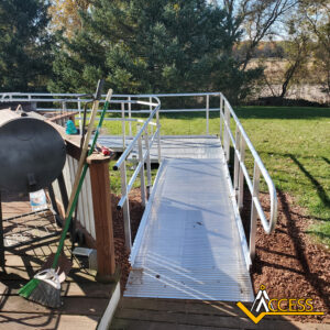 A metal wheelchair ramp with handrails leads from a wooden deck to a grassy yard. Cleaning tools, including a broom, lean against the deck. Trees and an open field are visible in the background. ACCESS logo in the corner.