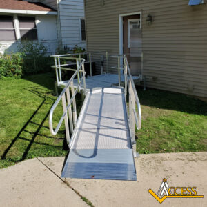 A metal wheelchair ramp with handrails leads from a concrete walkway to the back door of a beige house. The ramp is on a grassy lawn and features the Access company logo in the corner.