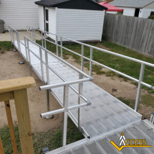 A metal wheelchair ramp with handrails leads from a wooden porch to a small white shed, crossing over grass and dirt. The ACCESS logo is in the bottom right corner.