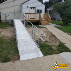 A metal wheelchair ramp with handrails leads from a sidewalk to the back door of a house. Nearby, there are steps with a wooden railing leading to a small porch. The Access logo is in the lower right corner.