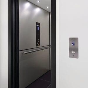 A modern open Zenith Elevator by Savaria features a stainless steel interior, horizontal handrail, and illuminated ceiling lights. The control panel and a lit call button are visible on the wall outside the elevator.