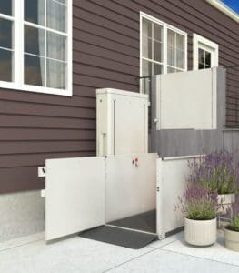 A VPL - 3100/3200 Series - Vertical Platform Lift by Bruno is installed outside a brown house near stairs and potted plants, providing reliable access to an elevated doorway.