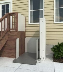 A Bruno VPL - 3100/3200 Series - Vertical Platform Lift by Bruno is installed beside a small wooden staircase, granting accessible entry to a yellow-sided house with white-trimmed windows. A green shrub is visible on the right.