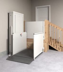 A VPL - 3100/3200 Series Vertical Platform Lift by Bruno is installed beside a wooden staircase, granting wheelchair access to an upper-level doorway in a carpeted indoor area.