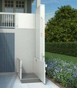 The VPL - 3100/3200 Series - Vertical Platform Lift by Bruno is installed outdoors beside a porch railing, granting wheelchair access from ground level to an elevated entryway, with hedges and flowering bushes in the background.