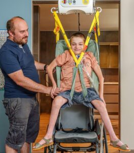 A caregiver assists a child using a SureHands portable ceiling lift and supportive sling to safely transfer into a wheelchair in a home setting.