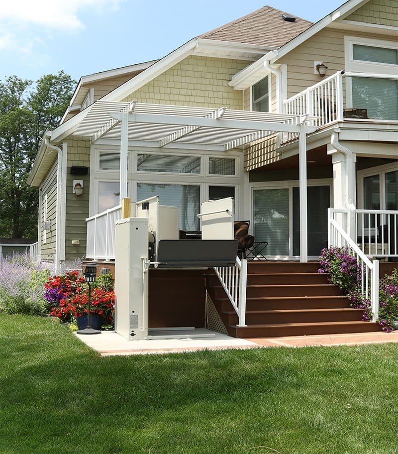A modern house features a raised wooden deck with white railings, a pergola, colorful flower pots, and a Bruno VPL-3100/3200 Series Vertical Platform Lift for easy yard-to-deck outdoor accessibility.