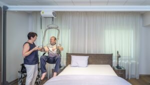 Caregiver assisting a patient using a SureHands ceiling lift with a body support sling in a bedroom, demonstrating a safe transfer from a chair to a bed using an overhead track system.