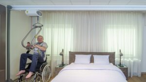 Patient using a SureHands ceiling lift with a body support sling to transfer safely between a wheelchair and a bed in a bedroom using an overhead track system.