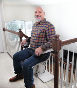 Person seated on a Bruno Elan SRE-5000 straight stair lift at the top of a residential staircase, demonstrating the swivel seat, seat belt, and folding footrest for safe and comfortable entry and exit.