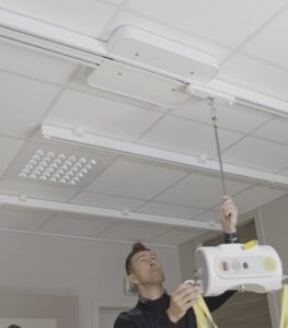Ceiling-mounted SureHands Manual Point patient lift system with a motor unit attached to an overhead track. A caregiver pulls the manual control cord to operate the lift in a clean indoor healthcare or home care setting, demonstrating safe and accessible patient transfer.