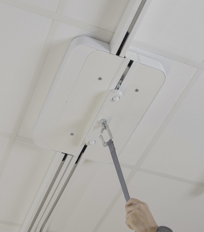 Ceiling-mounted SureHands Manual Point patient lift system with a motor unit attached to an overhead track. A caregiver pulls the manual control cord to operate the lift in a clean indoor healthcare or home care setting, demonstrating safe and accessible patient transfer.