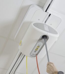 Ceiling-mounted SureHands Manual Point patient lift system with a motor unit attached to an overhead track. A caregiver pulls the manual control cord to operate the lift in a clean indoor healthcare or home care setting, demonstrating safe and accessible patient transfer.