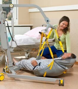 SureHands Model 1630 mobile patient lift lifting a patient from the floor using a full-body sling while a caregiver assists in a healthcare room, with a hospital bed in the background.