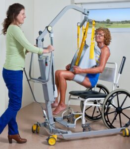 SureHands Model 1630 mobile patient lift transferring a patient into a wheelchair using a full-body sling while a caregiver assists in a healthcare setting.