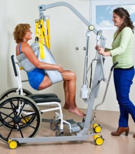 SureHands Model 1630 mobile patient lift transferring a patient into a wheelchair using a full-body sling while a caregiver assists in a healthcare setting.