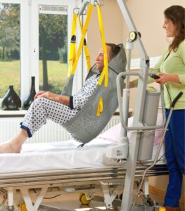 SureHands Model 1630 mobile patient lift lifting a patient from the floor using a full-body sling while a caregiver assists in a healthcare room, with a hospital bed in the background.