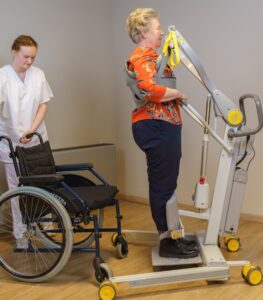 SureHands Stand Assist 2620 helping a patient safely transition from sitting to standing using a supportive standing sling while a caregiver assists, with a wheelchair positioned nearby for transfer.
