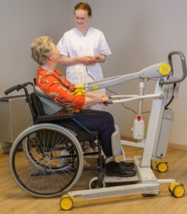 SureHands Stand Assist 2620 transporting a patient while seated in a wheelchair using a supportive standing sling, with a caregiver operating the powered lift via handheld remote in a home care setting.